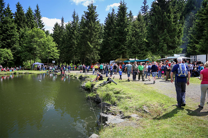 Menschenansammlung bei einem Fest am Ufer eines Waldteiches. Im Hintergrund Verkaufsstände und Zelte, umgeben von hohen Tannen. Sonniges Wetter.
