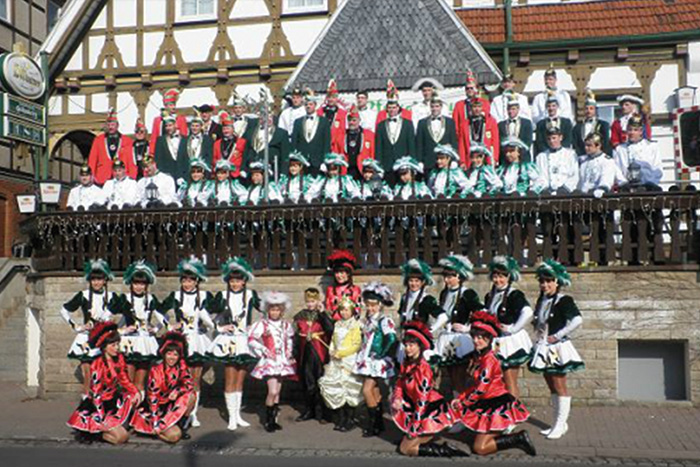 Gruppenfoto einer Karnevalsgesellschaft vor einem Fachwerkgebäude. Männer in festlichen Uniformen und Narrenkappen stehen auf einer Empore, davor Tanzgarden in bunten Kostümen.
