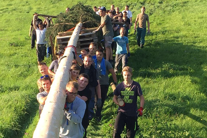 Gruppe junger und erwachsener Männer trägt gemeinsam einen langen Baumstamm mit Tannenspitze über eine grüne Wiese. Traditioneller Brauch, aufstellen einer Kirmestanne.