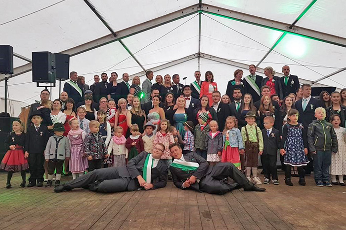 Gruppenfoto in einem Festzelt mit Erwachsenen und Kindern in festlicher Kleidung, darunter traditionelle Trachten. Zwei Männer in Anzügen mit Schärpen liegen lachend im Vordergrund auf dem Boden.