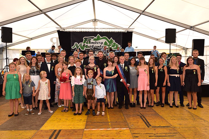 Gruppenfoto in einem Festzelt vor der Bühne der „Famberg Musikanten“. Erwachsene und Kinder in festlicher Kleidung, einige in Tracht, stehen zusammen und lächeln in die Kamera.
