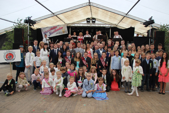 Großes Gruppenfoto in einem Festzelt vor einer Bühne mit Musikanten. Erwachsene und viele Kinder in festlicher Kleidung, teilweise in Tracht, stehen und sitzen gemeinsam vor der Kapelle.