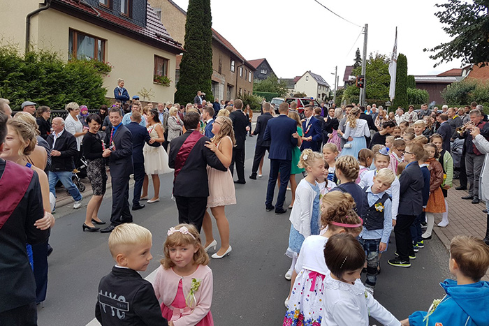 Straßenszene eines Dorffestes mit tanzenden Paaren in festlicher Kleidung und vielen Zuschauern. Auch Kinder nehmen am Tanz teil, im Hintergrund stehen zahlreiche Gäste am Straßenrand.
