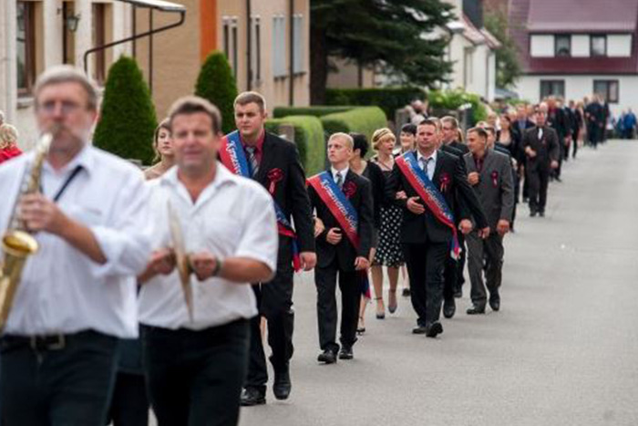 Festumzug mit Musikern und Teilnehmern in Anzügen, einige mit blauen Schärpen und roten Blumenansteckern. Die Gruppe marschiert in Reihen eine Dorfstraße entlang.