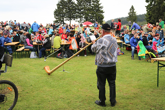 Mann spielt Alphorn auf einer Wiese vor zahlreichen Besuchern, die an Biertischen sitzen. Veranstaltung im Freien mit vielen Menschen in wetterfester Kleidung.