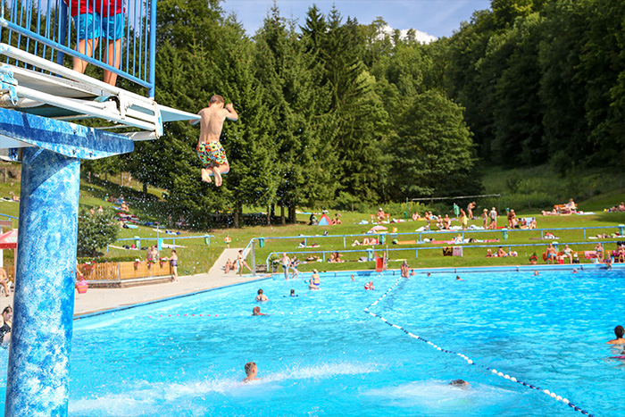 Blick auf das Schwimmbecken, im Vordergrund der Sprungturm von dem ein Kind springt