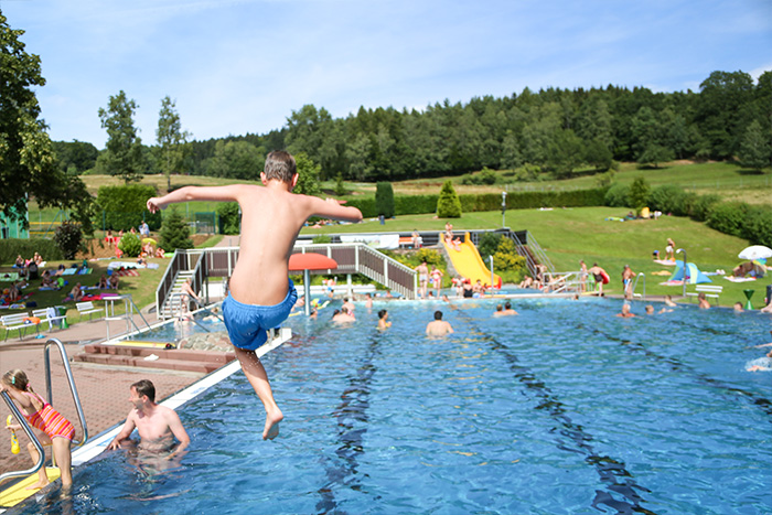 Kind springt in das Schwimmbecken, im Hintergrund ziehen Schwimmende ihre Bahnen