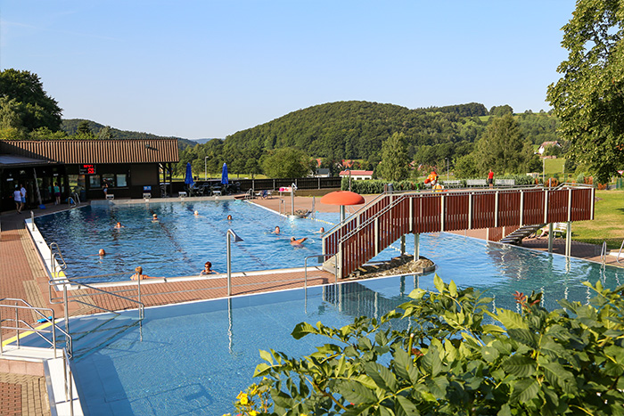 Außenaufnahme des Schwimmbads Struth-Helmershof mit Becken, Brücke und umgebender Landschaft