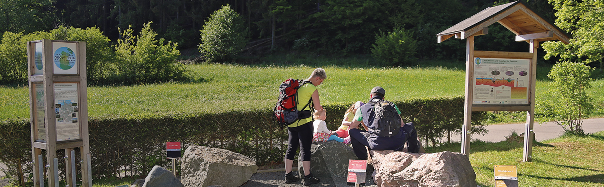Familie sitz auf ein paar Felsen umgeben von 2 Schautafeln im Geopark