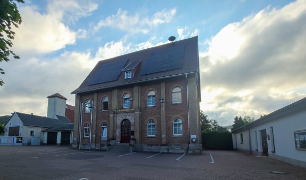 Backsteingebäude der Gemeindeverwaltung mit Solarpanels auf dem Dach, fotografiert bei bewölktem Himmel in den frühen Abendstunden.