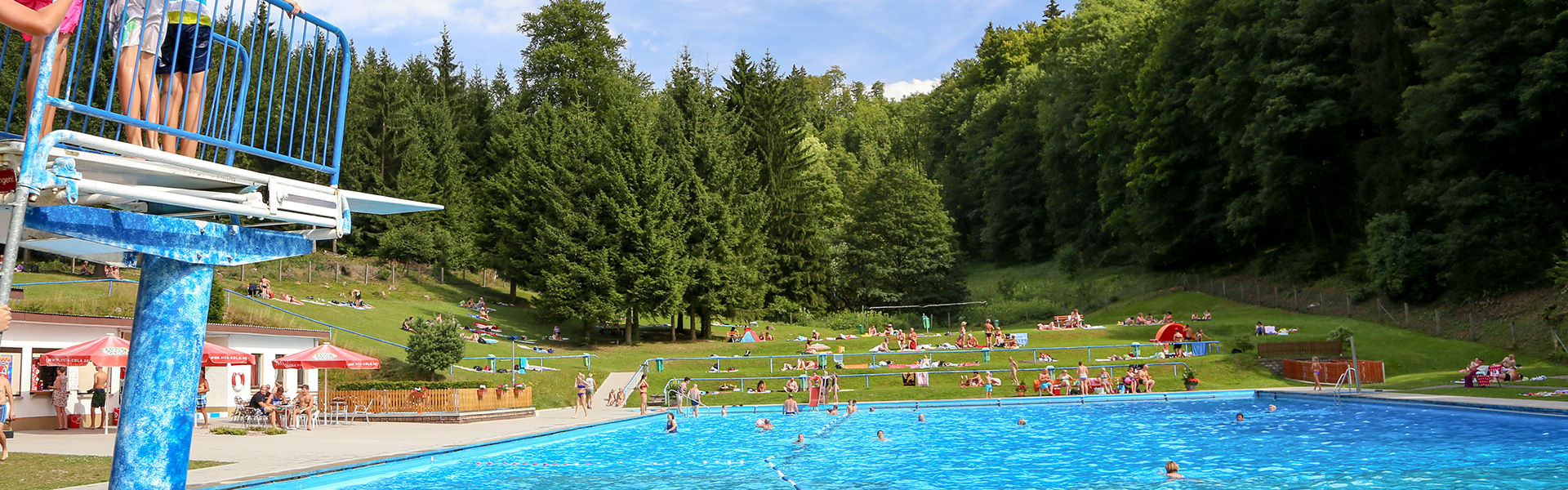 Bildausschnitt: Blick ins Schwimmbad, Sprungturm und Schwimmbecken mit Besuchern