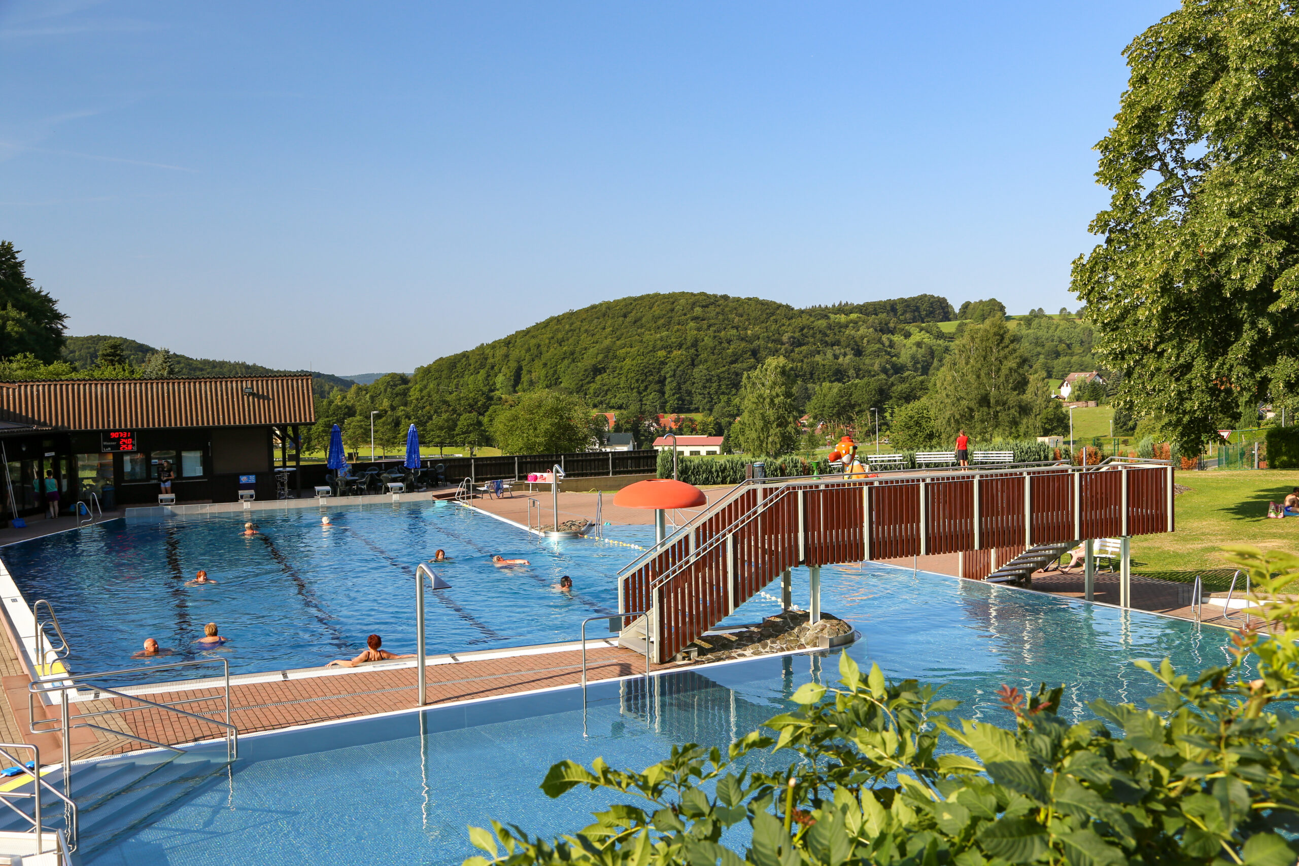 Außenaufnahme des Schwimmbads Struth-Helmershof mit Becken, Brücke und umgebender Landschaft