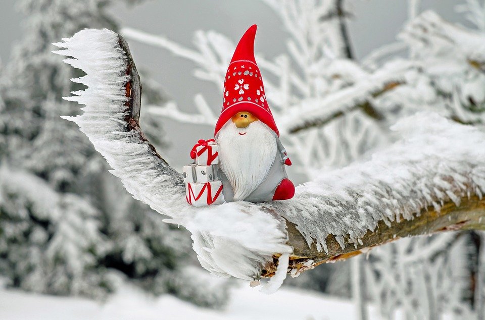 Weihnachtlicher Wichtel mit roter Mütze sitzt auf einem schneebedeckten Ast und hält zwei kleine Geschenke. Im Hintergrund verschneite Winterlandschaft.
